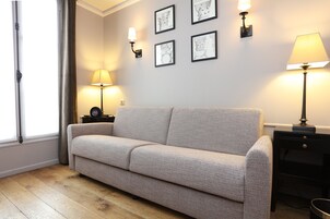 A living room with a beige sofa, a side table with a lamp, and wall-mounted picture frames.