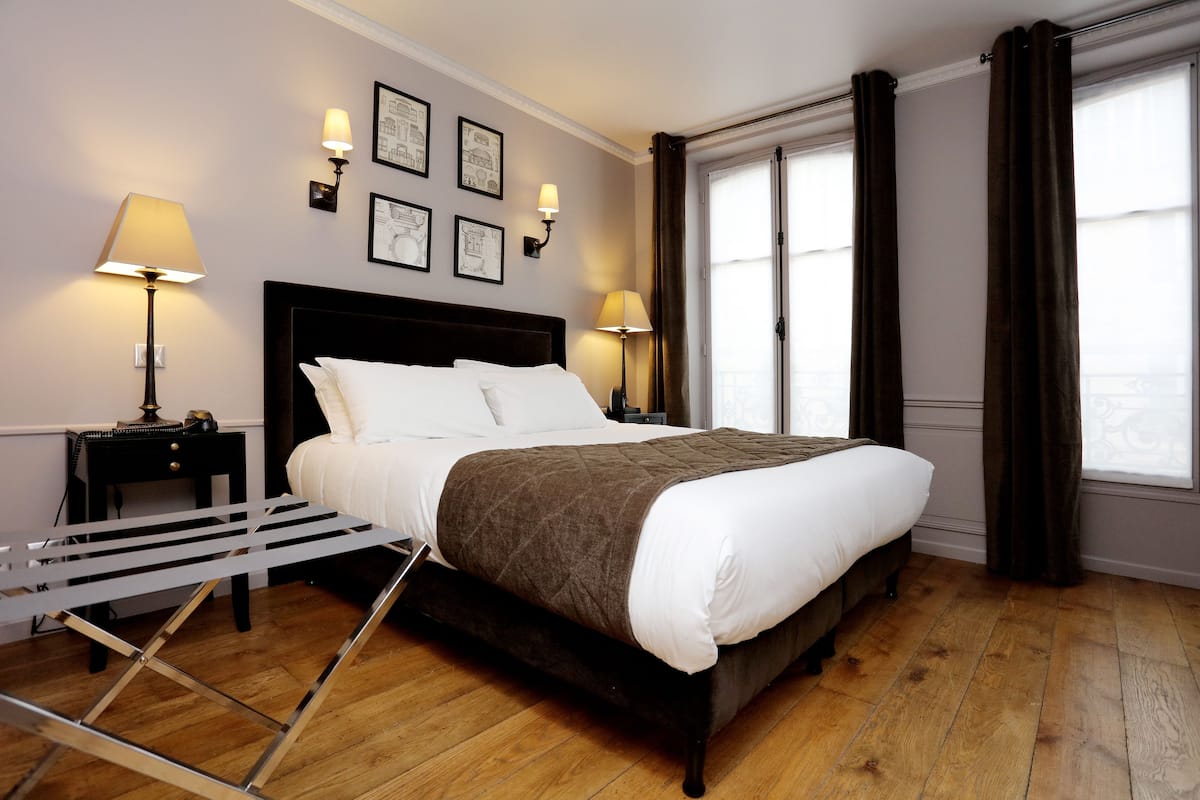 Una cama bien hecha con un cabecero oscuro, dos lámparas de noche y una ventana con cortinas.