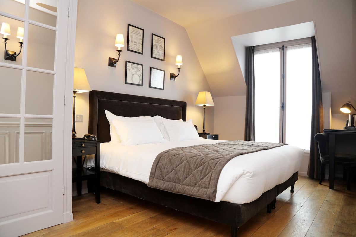 Una cama bien hecha con un cabecero oscuro, dos mesitas de noche con lámparas, un ventanal con cortinas y cuadros en la pared.