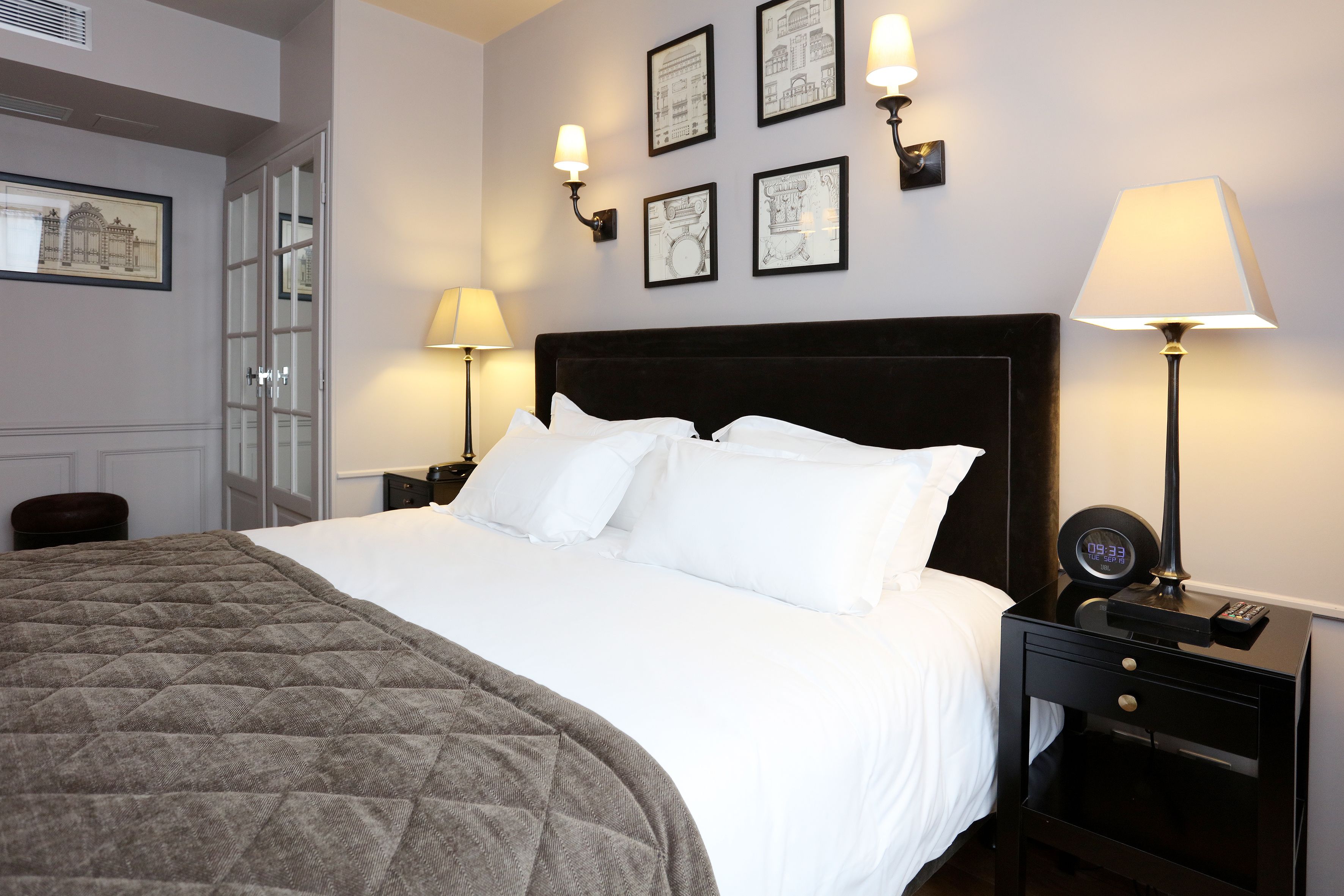 A neatly made bed with white linens and a quilted blanket, a bedside table with a lamp, and framed pictures on the wall.