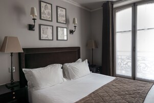 A neatly made bed with white linens and a brown quilt, flanked by two bedside tables with lamps, in a room with framed artwork on the wall and a large window with a view.
