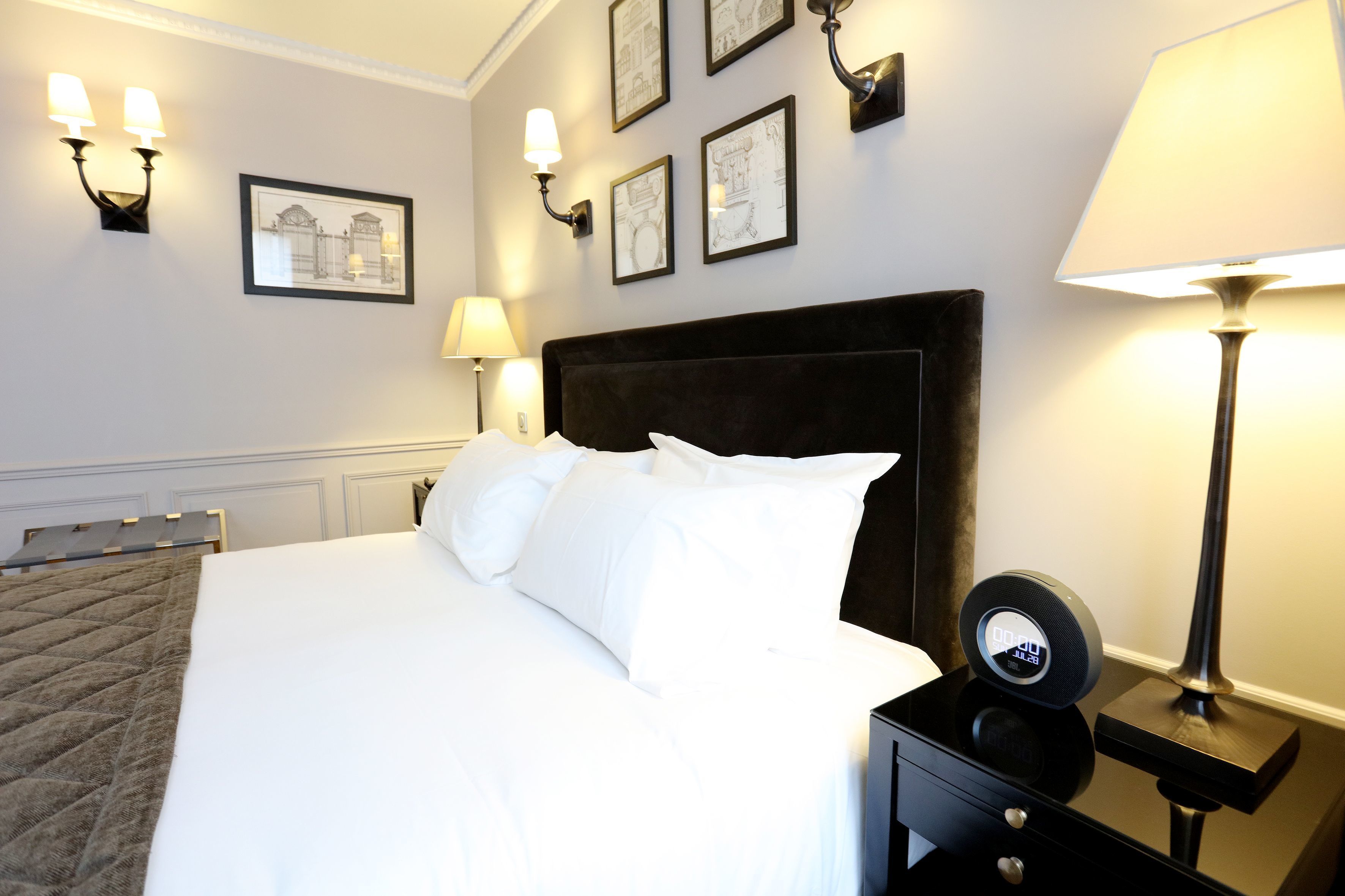 Una cama bien hecha con sábanas blancas, un reloj digital en una mesita de noche y lámparas fijadas a la pared.