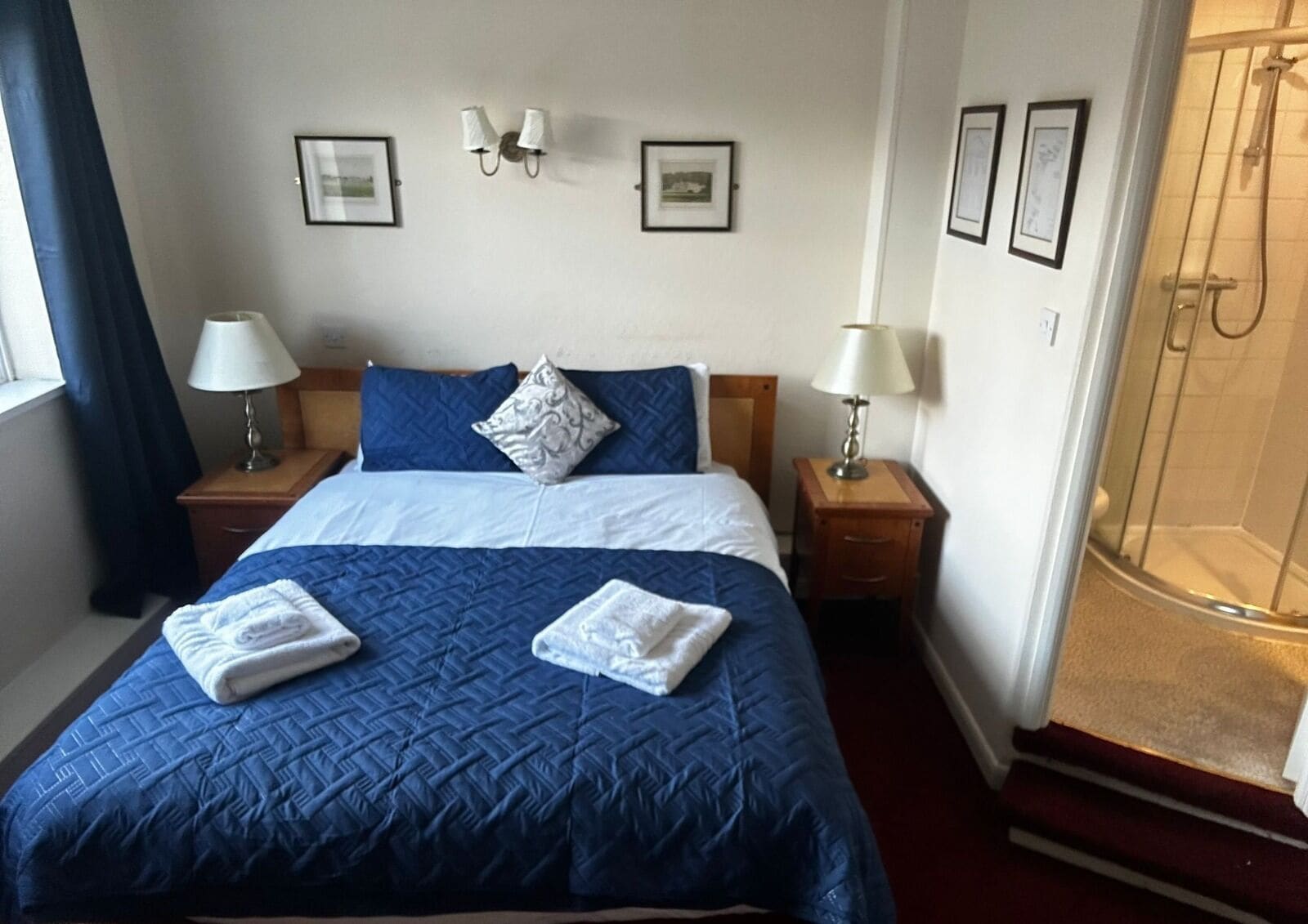 Habitación de hotel con una cama grande, dos mesitas de noche con lámparas, zona de ducha y cuadros en las paredes.