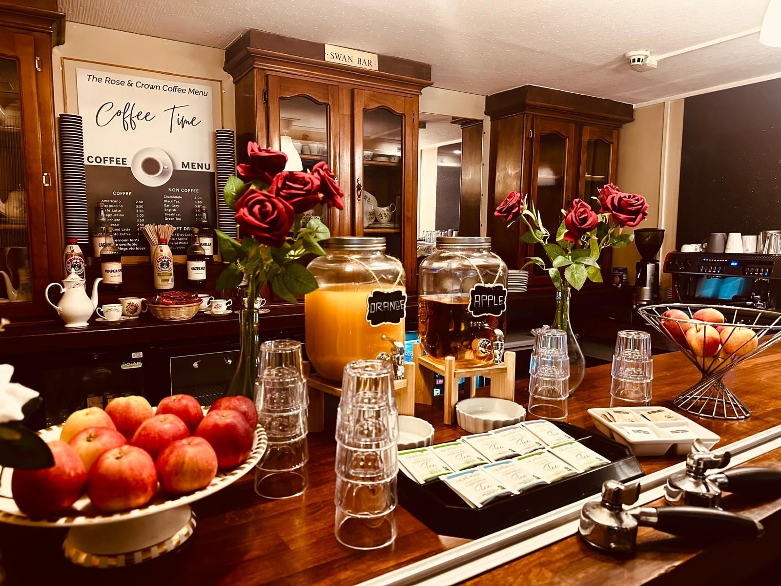 Una estación de café con un menú, frascos etiquetados con bebidas y un bol de manzanas.