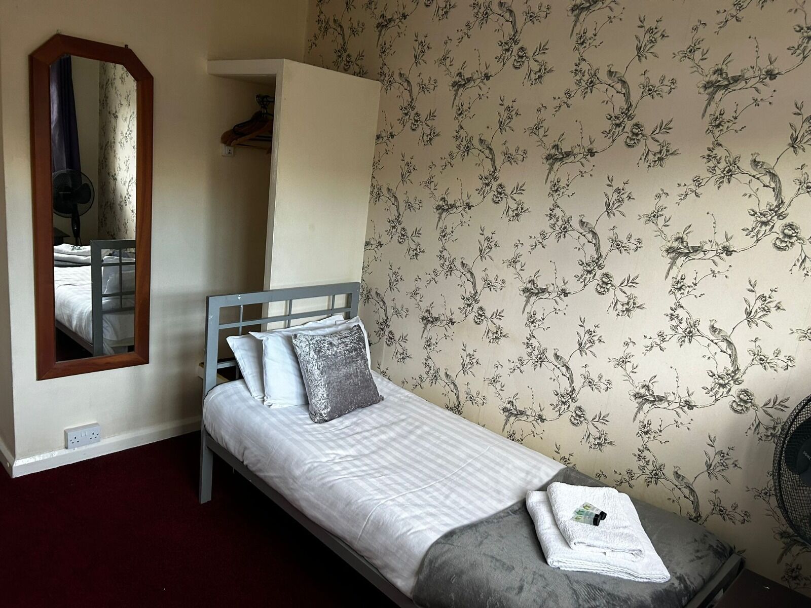 A single bed with a grey headboard, white bedding, and a grey pillow. A mirror is mounted on the wall to the left.