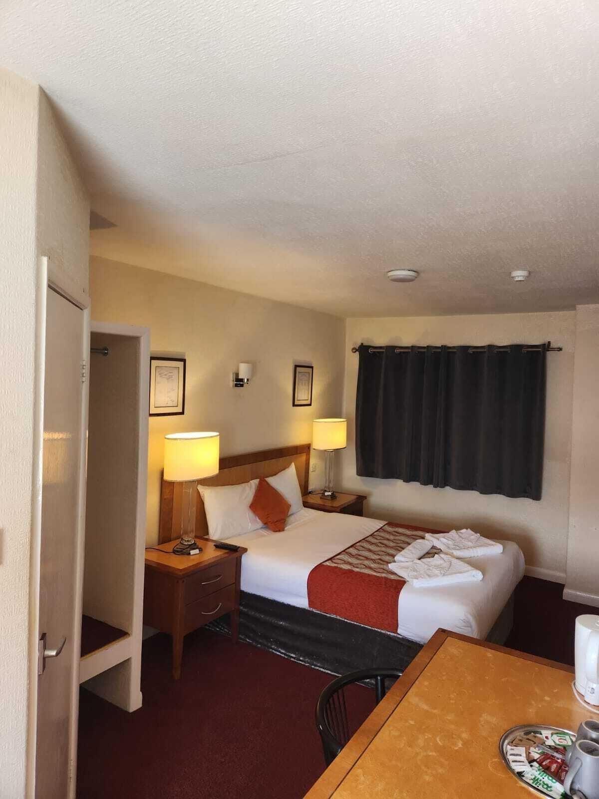 A hotel room with a bed, two bedside lamps, a nightstand, a table with a coffee maker, and a window with curtains.