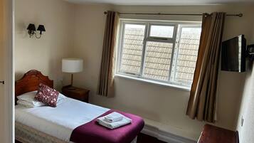 A neatly made bed with a white and purple bedspread, a wooden headboard, a bedside lamp, and a window with curtains.