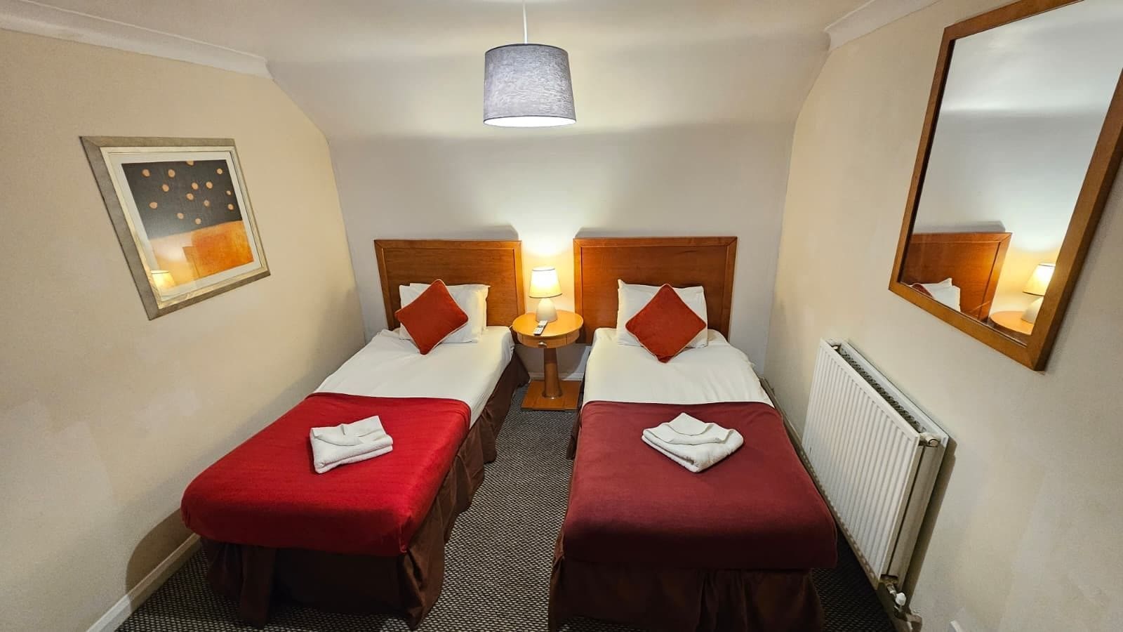 A hotel room with two beds, a nightstand, a lamp, a mirror, and a radiator.