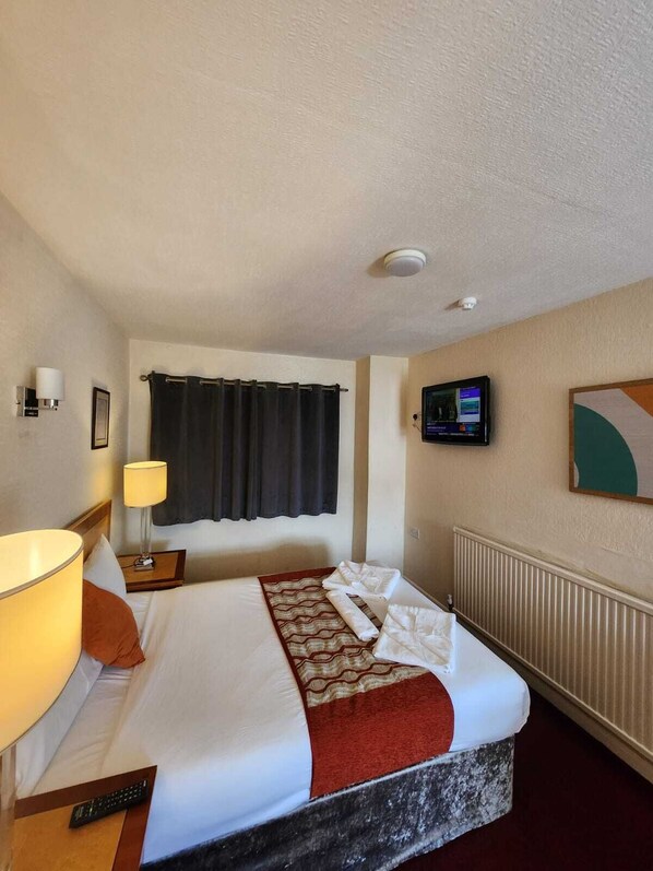 A hotel room with a bed, a nightstand, a lamp, a television, and a wall-mounted control panel.