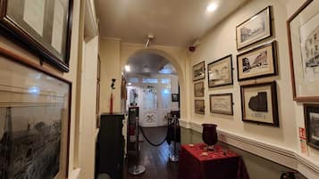 A corridor with framed pictures on the walls, a red table with a vase, and a fire extinguisher.