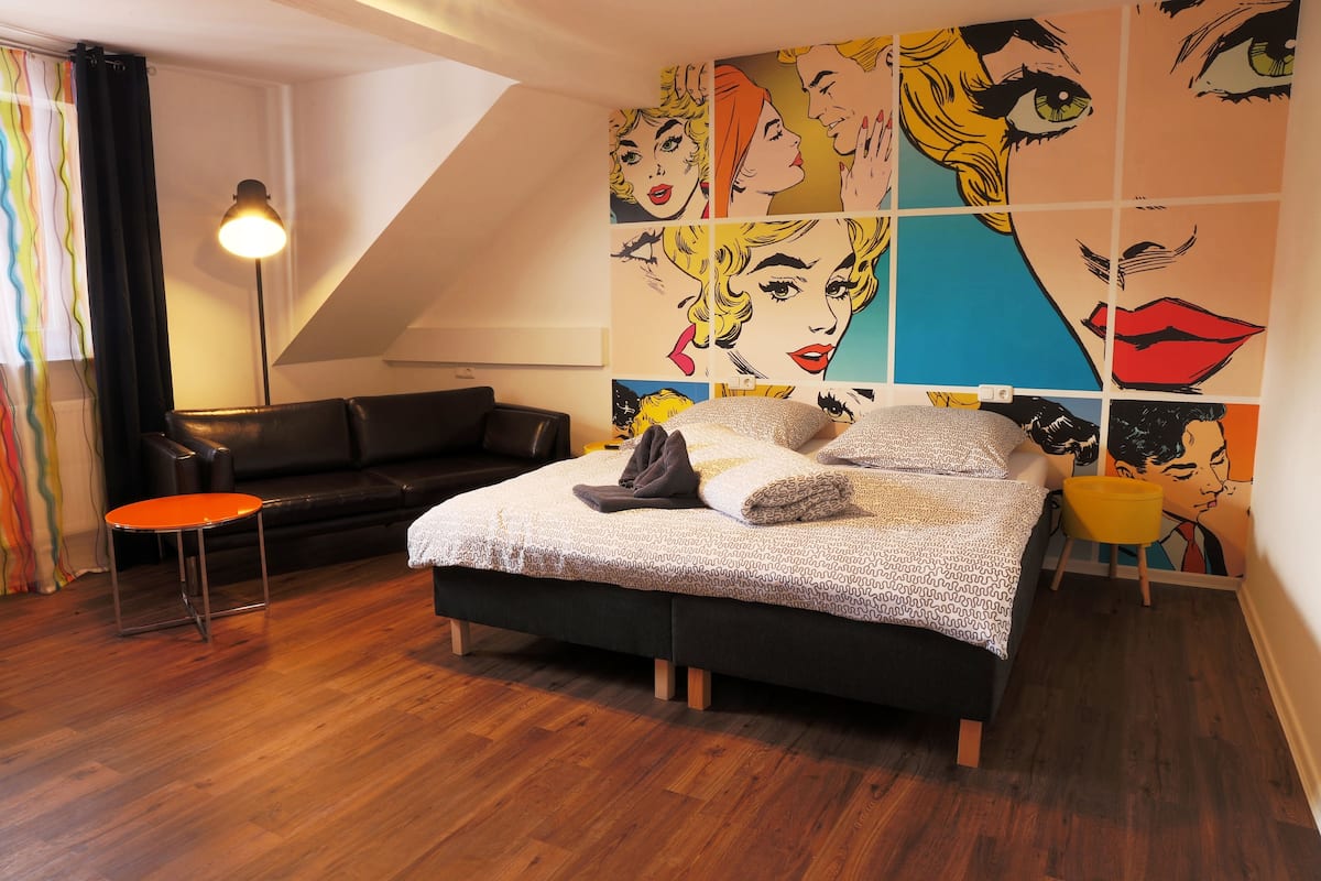 Een slaapkamer met een bed, een stoel, een lamp en een muur met pop-art prints.