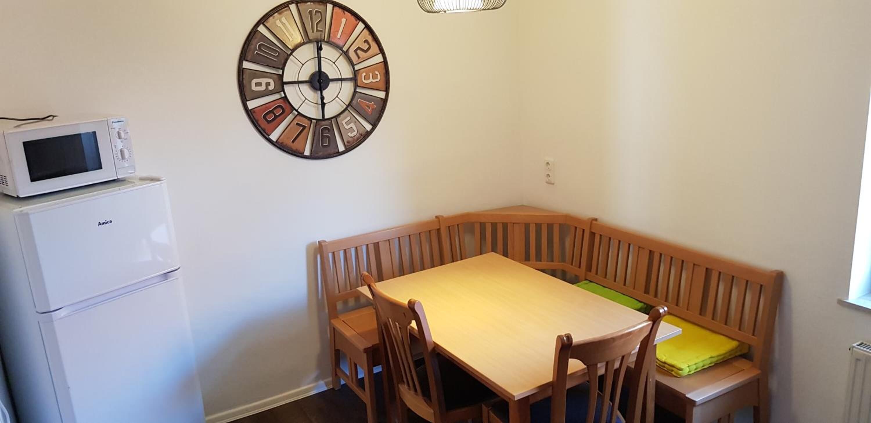 A small room with a wooden table and benches, a wall clock, a microwave, and a refrigerator.