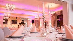 Une salle à manger aménagée avec élégance, où les tables sont dressées pour accueillir plusieurs convives, avec des nappes blanches, de la verrerie raffinée et des chandeliers.