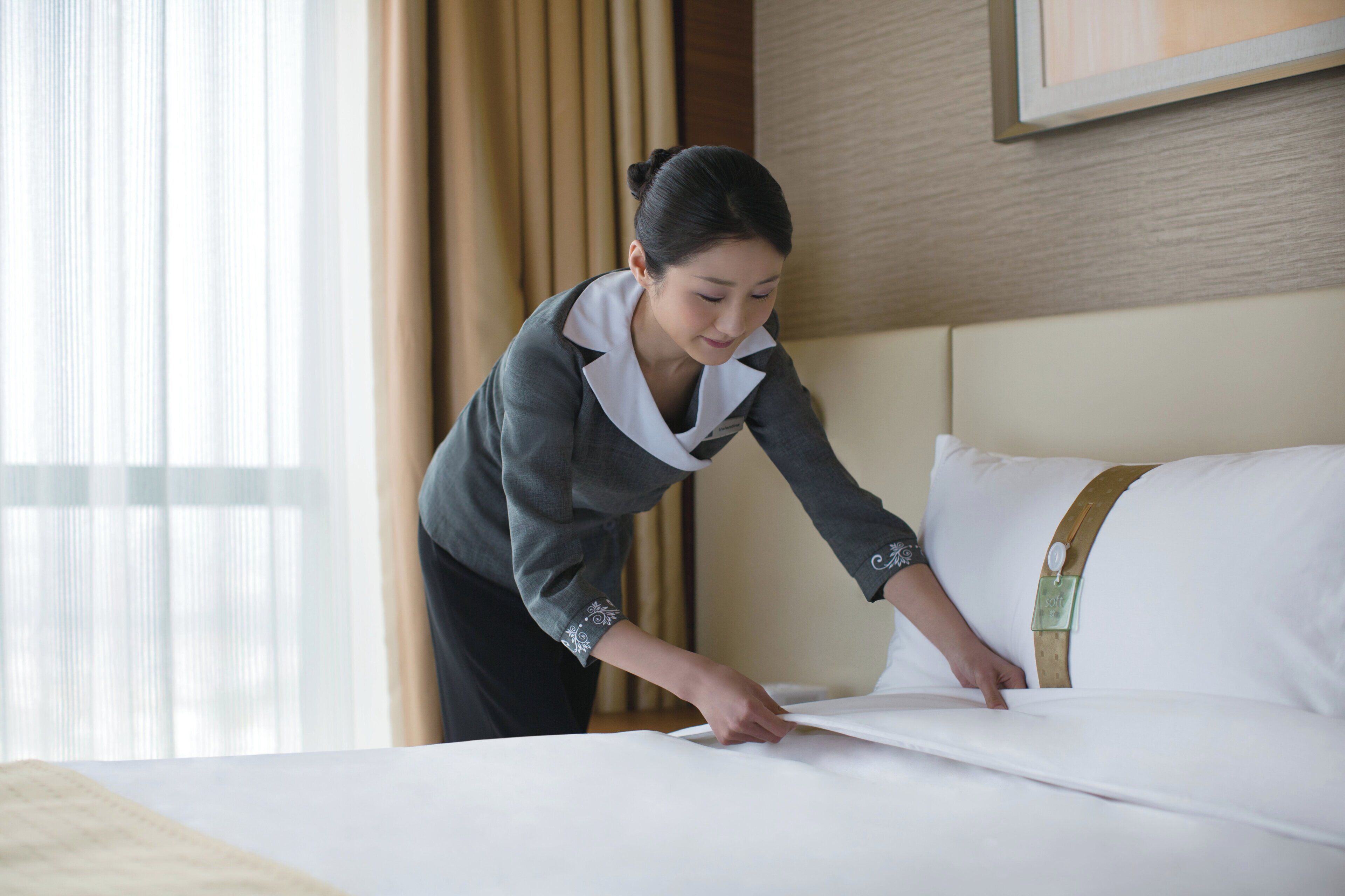 A hotel room with a neatly made bed, a pillow with a decorative strap, and a window with curtains.