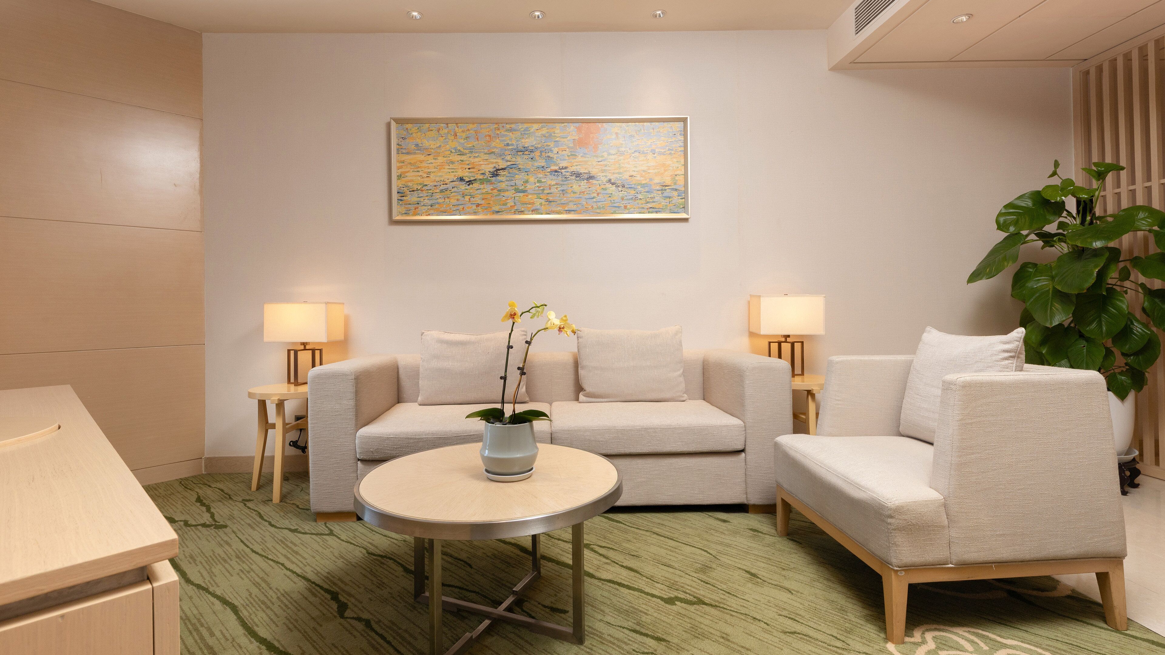 A modern living room with a sofa, armchair, round table, two lamps, a painting, and a potted plant.