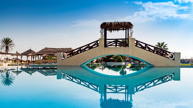 Une piscine d’eau cristalline, avec un pont en bois et, au fond, des constructions aux toits de chaume.