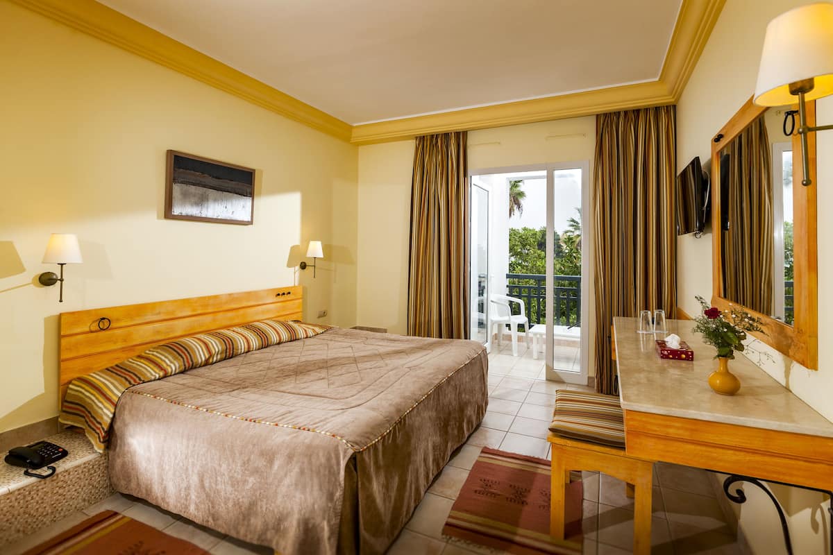 A hotel room with a bed, bedside lamps, a desk with a vase, and a view of the outdoors.