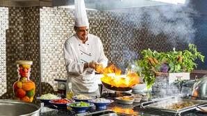 Un chef cuisinant avec un wok sur une cuisinière, entouré de divers ingrédients et devant un mur recouvert de carreaux.