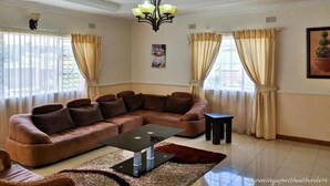 A living room with a brown leather sofa, a glass coffee table, and a chandelier. - Home Up Guest House (Blantyre)