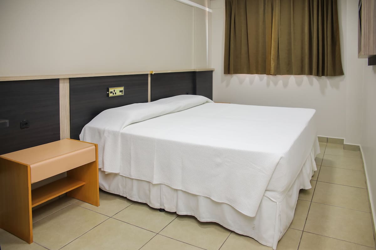 A neatly made bed with a white bedspread, a wooden nightstand, and a window with curtains.