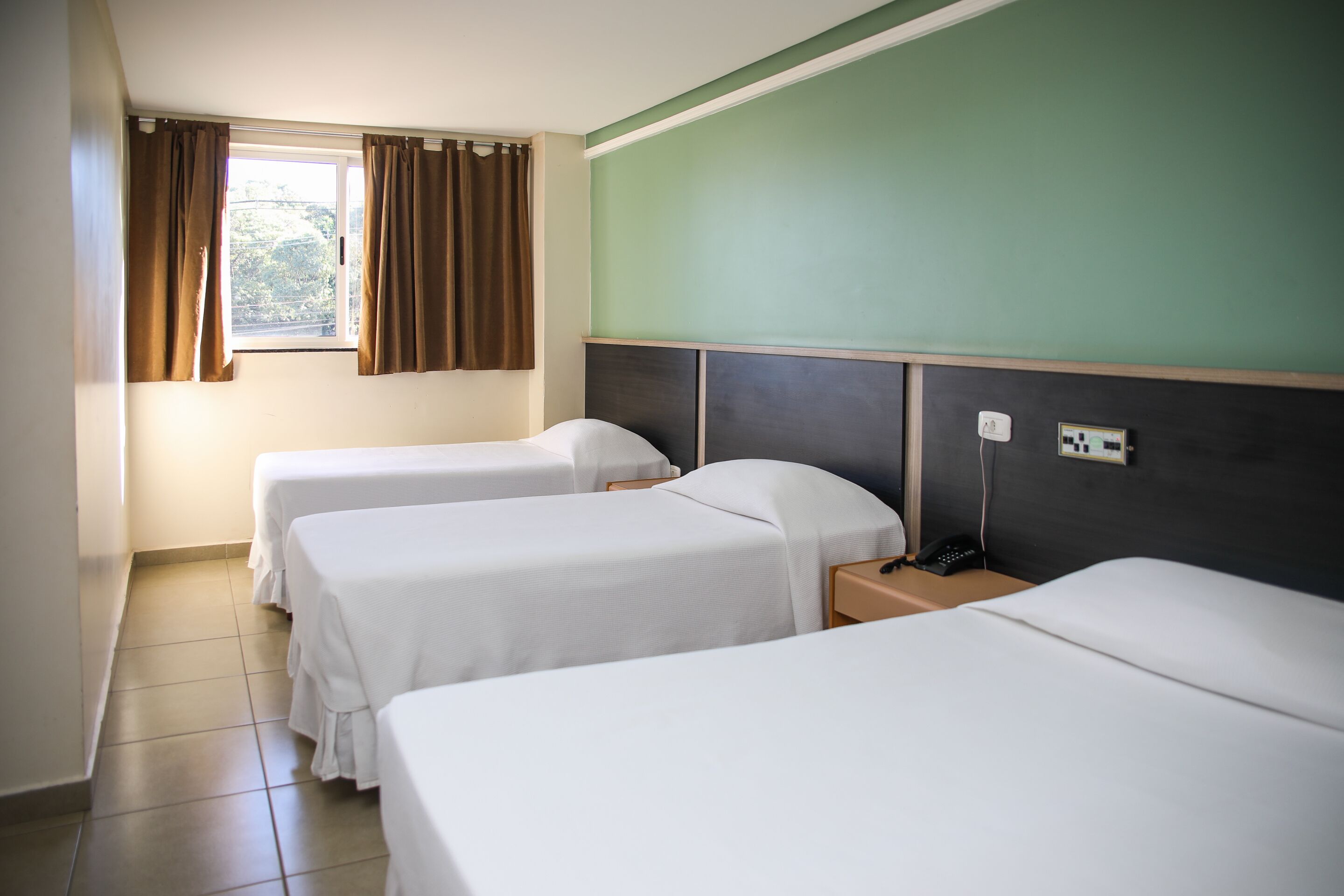 Ett hotellrum med två separata sängar, ett fönster med gardiner, ett nattduksbord med en telefon och en väggmonterad ljusströmbrytare.