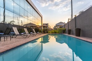 A swimming pool with lounge chairs and umbrellas, surrounded by a modern building with large windows. - Fallsway Suites (Lusaka)