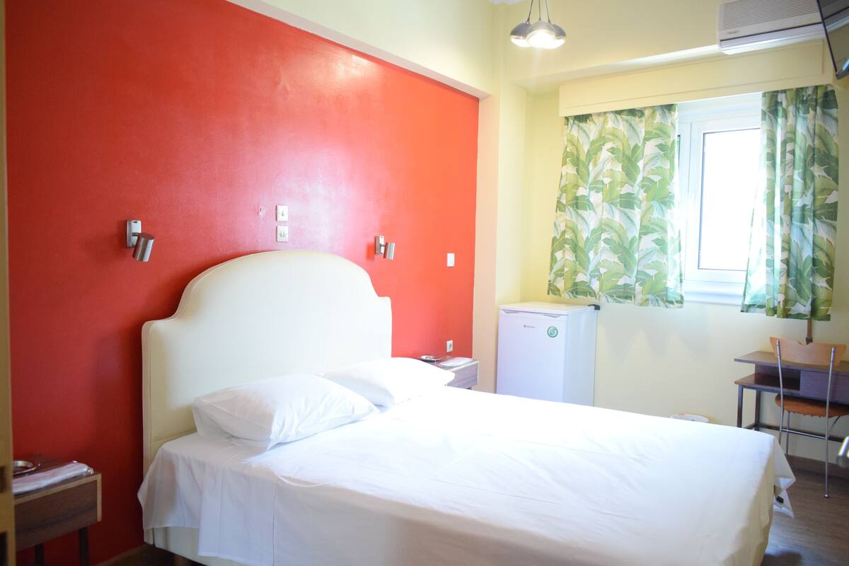A hotel room with a bed, a small desk, a refrigerator, and a window with curtains.