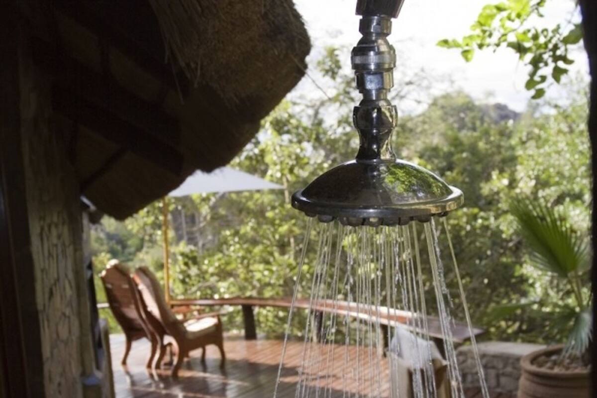Une pomme de douche laissant couler l’eau, une chaise en bois et un mur de pierre, le tout dans un décor de jardin.