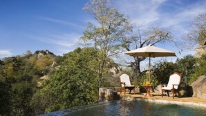 An outdoor swimming pool with two chairs and a table, surrounded by trees and rocks. - Sango Wildlife Lodge (Save Valley Conservancy)