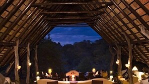 An outdoor dining area with a thatched roof, tables set for a meal, and candles providing ambient lighting. - Sango Wildlife Lodge (Save Valley Conservancy)