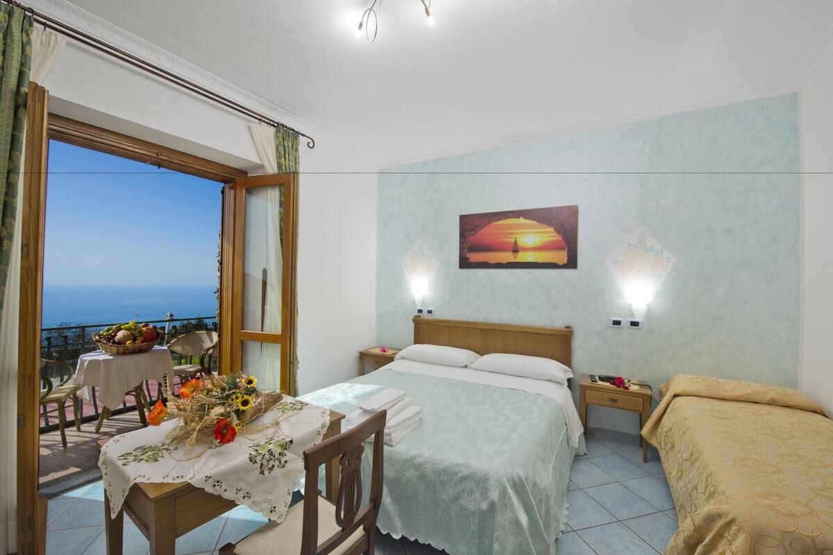 Une chambre à coucher comprenant un lit, une table de chevet, une commode, une fenêtre avec des rideaux, un balcon avec une table et des chaises, et un tableau accroché au mur.