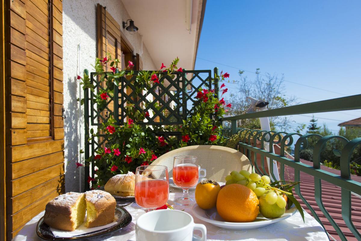 Une table dressée avec un gâteau, des fruits et des boissons sur un balcon, le tout agrémenté d’une treille en arrière-plan et de fleurs.