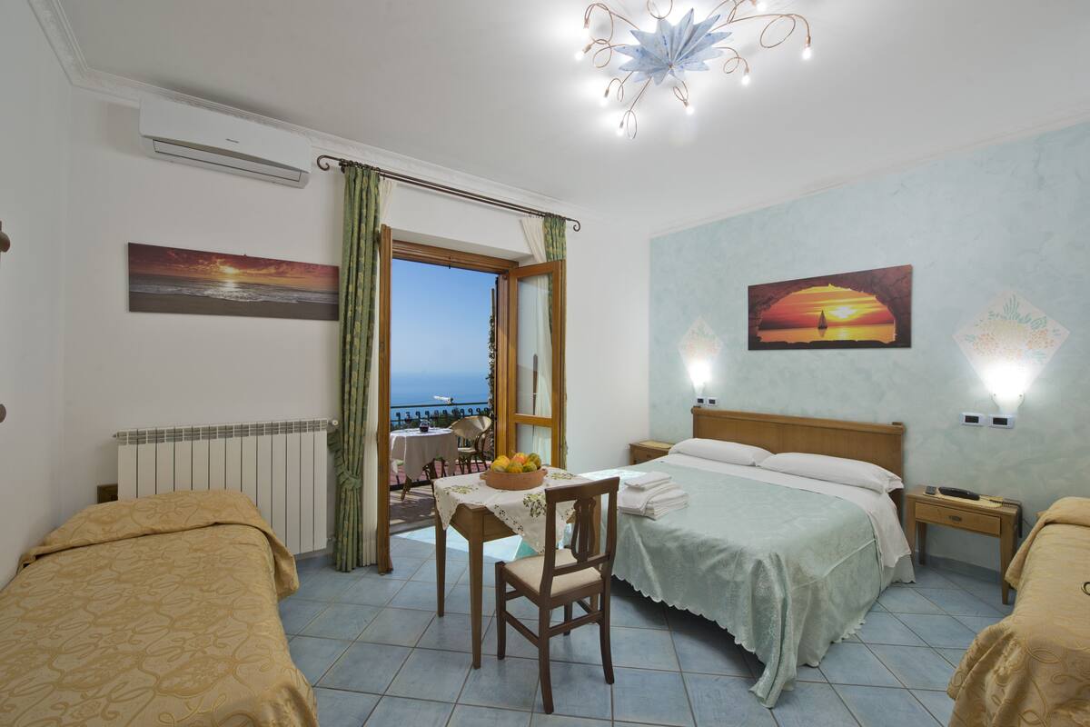Une chambre d’hôtel avec un lit, une table sur laquelle se trouve un panier de fruits, une chaise, une table de chevet et une vue sur la mer par une porte ouverte.