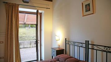 A bedroom with a bed, a nightstand, a wall-mounted lamp, a door leading to an outdoor area, and a framed picture on the wall.