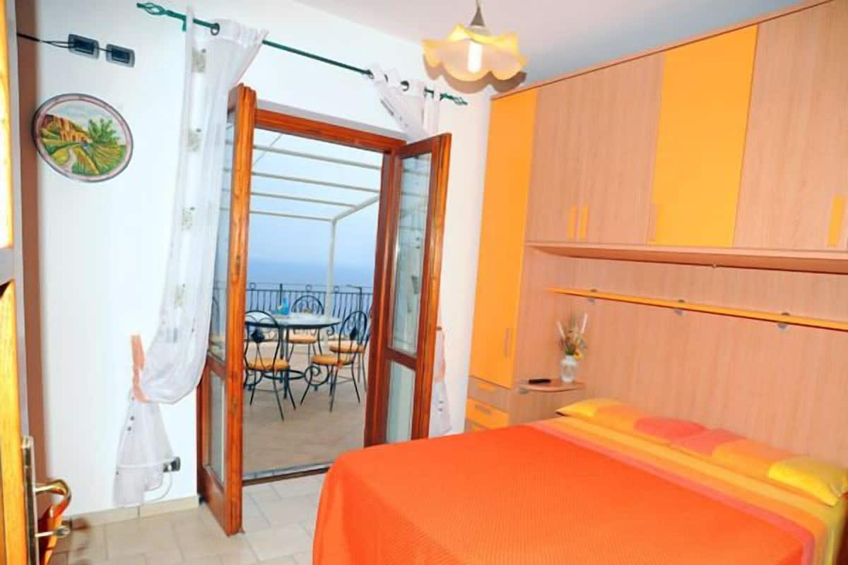 A bedroom with a bed, a balcony with a table and chairs, and a view of the sea.