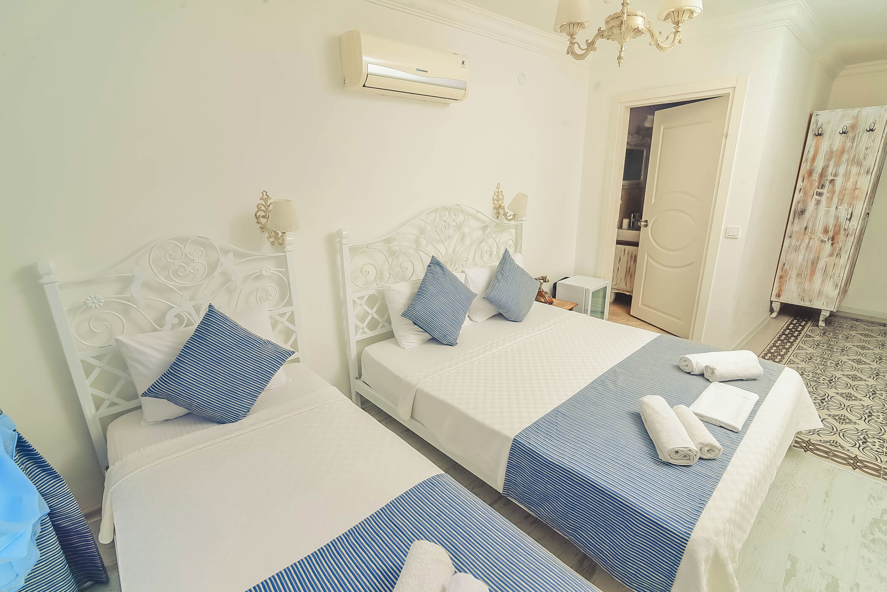 A hotel room with two beds, each with a white headboard and blue bedding. There is an air conditioning unit on the wall, a chandelier, and a patterned rug on the floor.