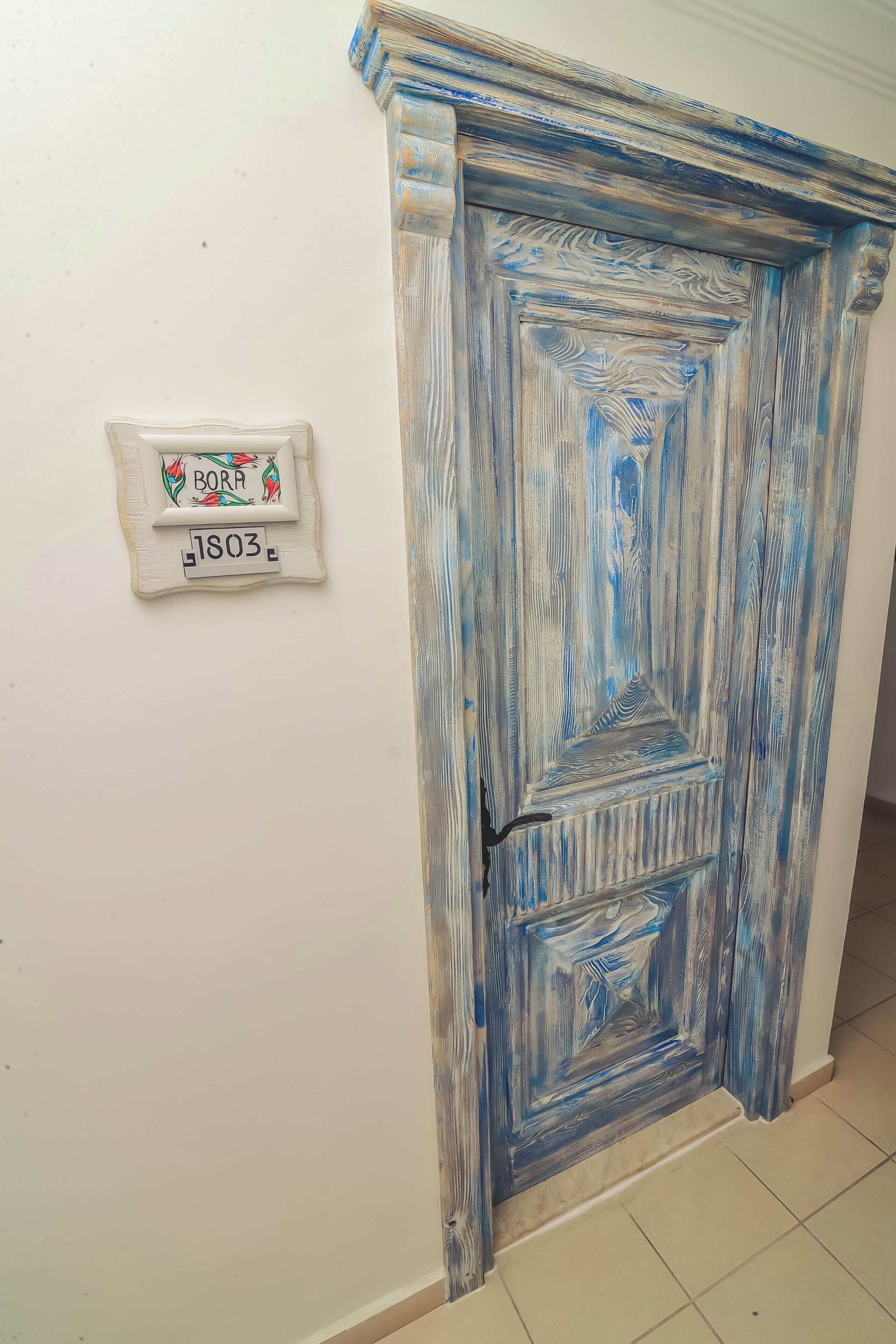 A blue-painted door with a decorative mold trim and a plaque with the name "BORA" and the number "1803".