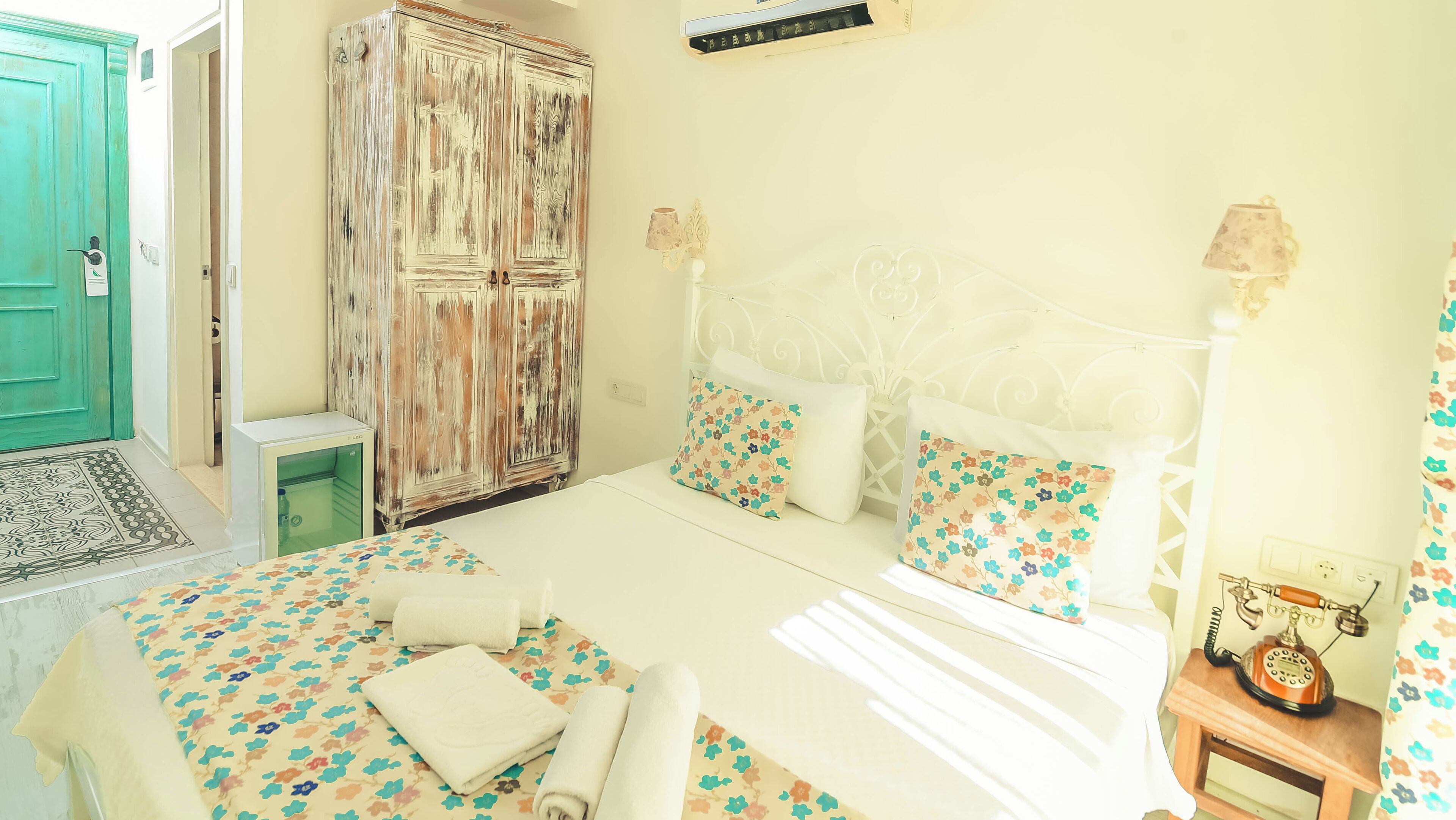 Un dormitorio con cama, almohadas, una mesita de noche con teléfono, un armario y una puerta.