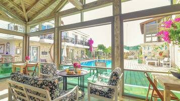 A covered outdoor seating area with a pool view, wooden furniture, and a stone wall.