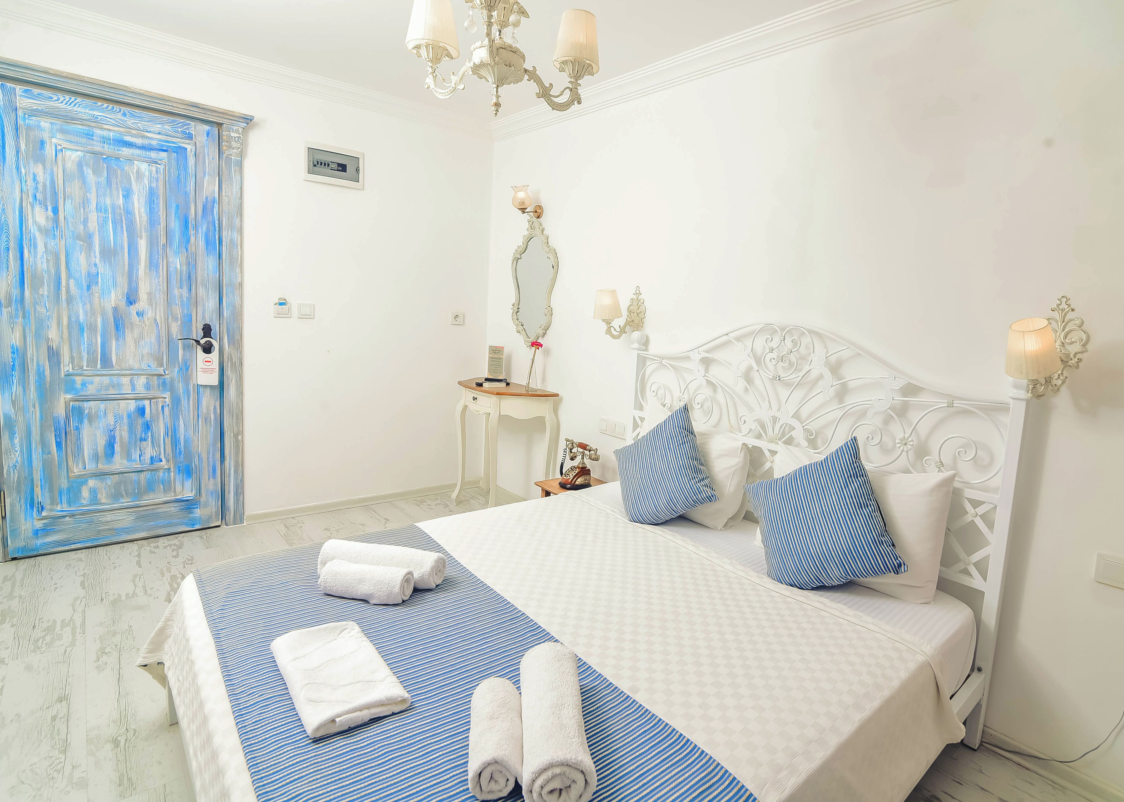 Un dormitorio con cama blanca, colcha a rayas azul y blanco, y almohadas a juego. Hay una puerta azul, una mesita con espejo, y una lámpara de pared.