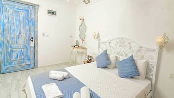 Un dormitorio con cama blanca, colcha a rayas azul y blanco, y almohadas a juego. Hay una puerta azul, una mesita con espejo, y una lámpara de pared.