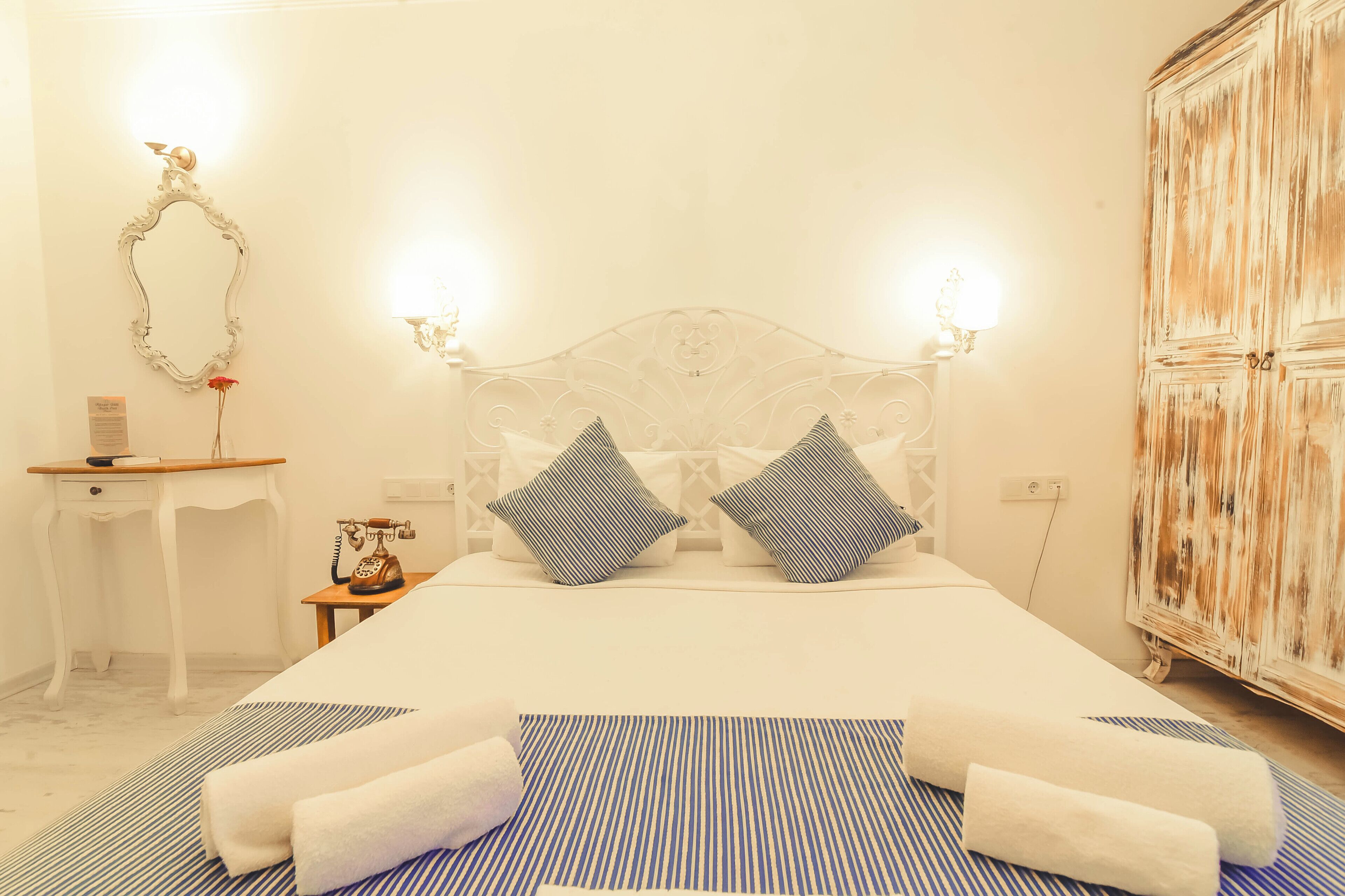 A neatly arranged bedroom with a large bed, two pillows, a bedside table with a telephone, a mirror, and a wardrobe.