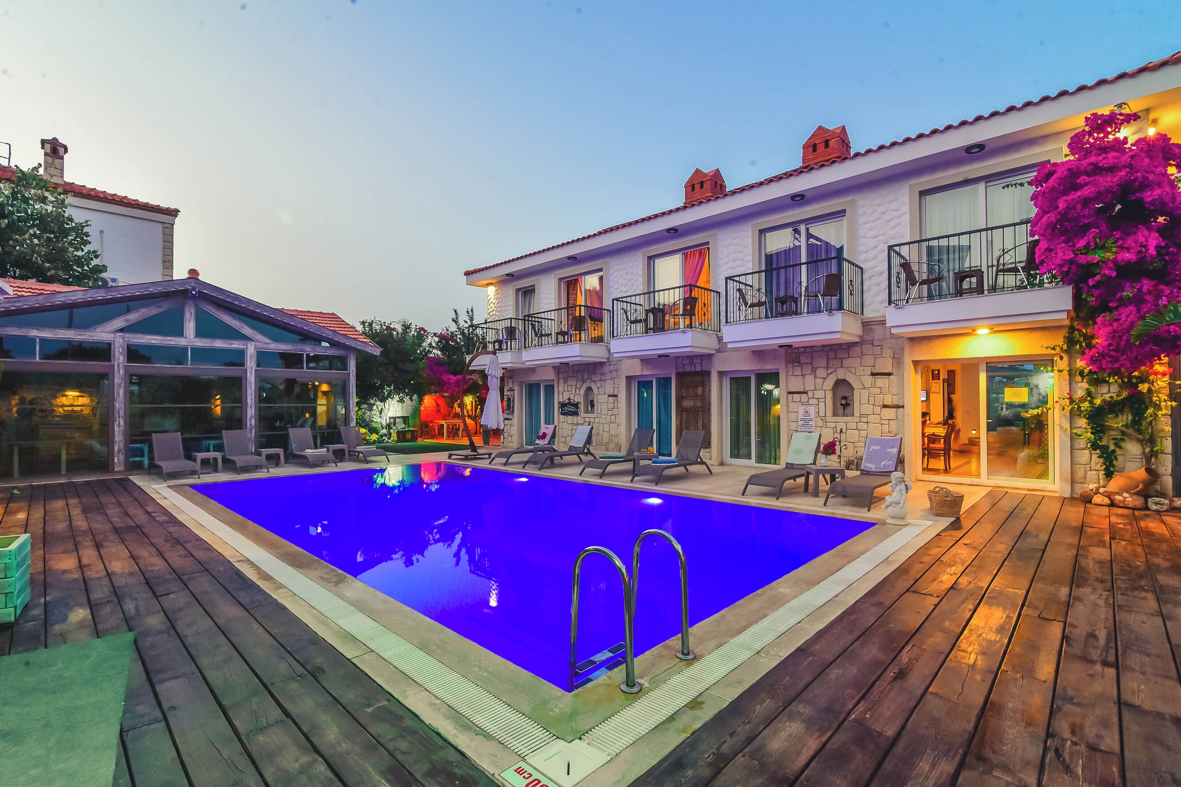 A swimming pool with a wooden deck, a covered area, and a building with balconies and outdoor seating.