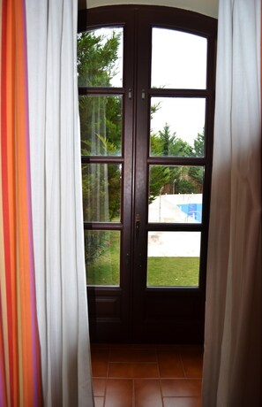 A dark wooden door with a glass panel, white curtains, and a view of a swimming pool and greenery outside. - Hotel El Carmen (Puente Genil)
