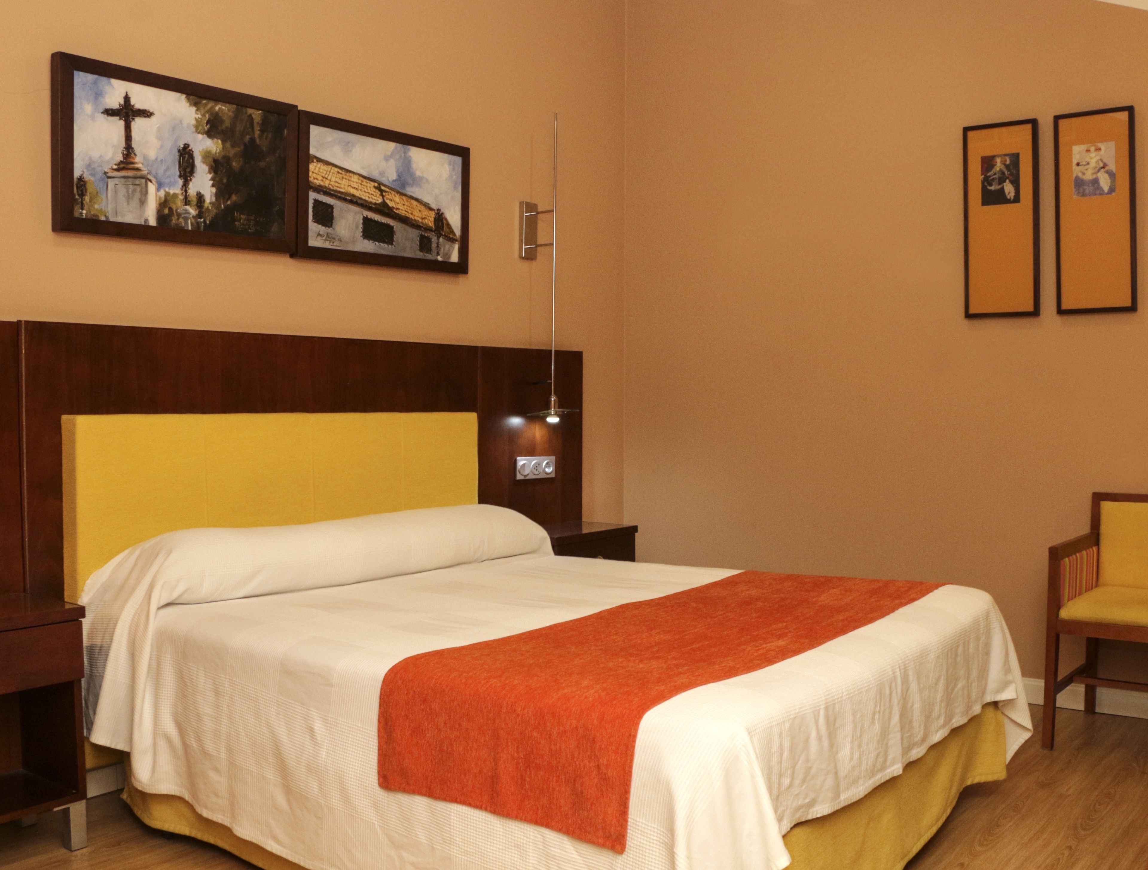 A hotel room with a bed, a nightstand, a chair, and two framed pictures on the wall.