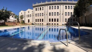 A swimming pool with a clear blue water surface, surrounded by a white building with multiple windows and a balcony. - Hotel El Carmen (Puente Genil)