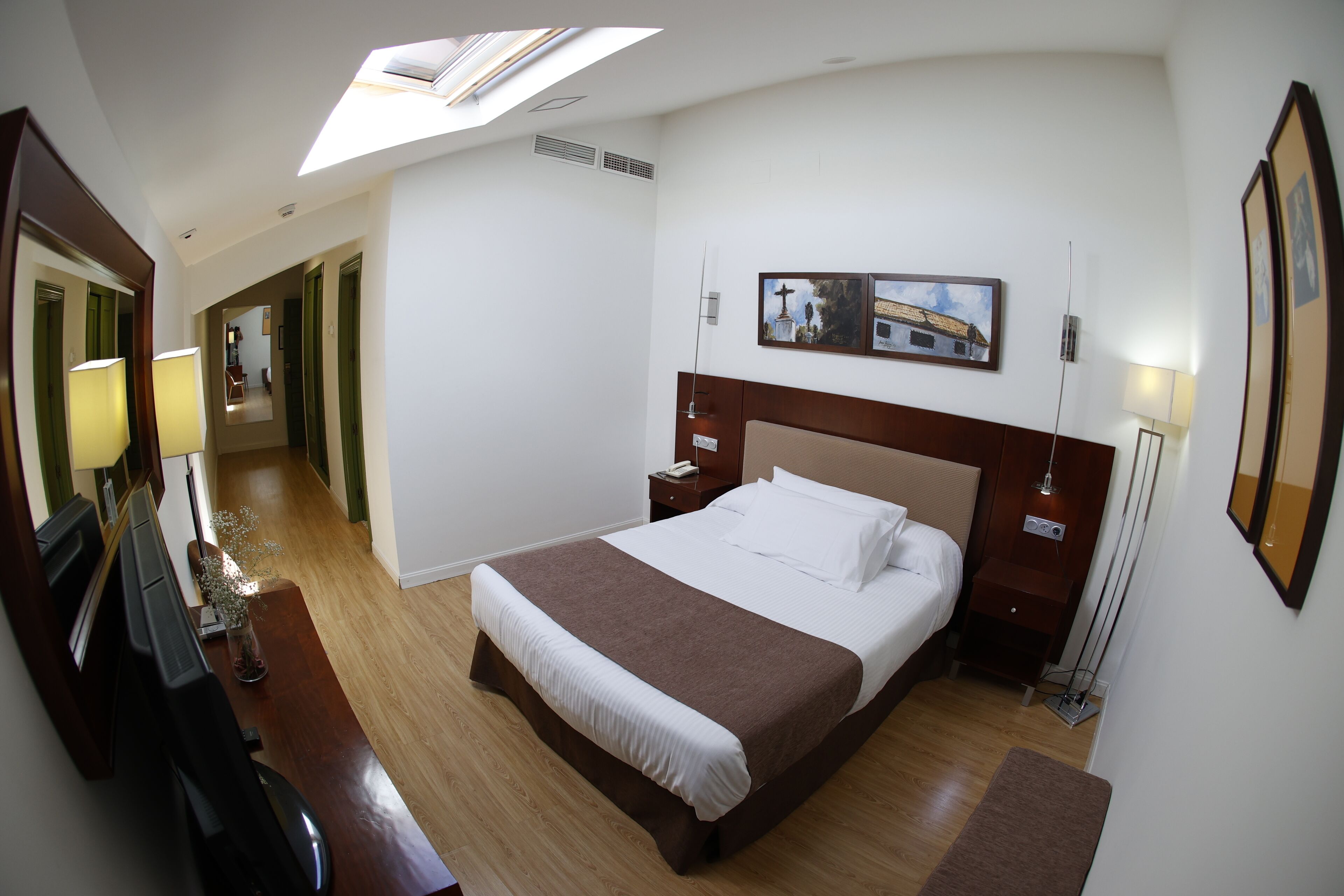 A hotel room with a large bed, a desk with a chair, a standing lamp, and a skylight.