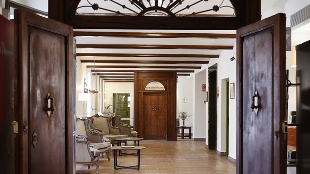 A hallway with wooden doors, a bench, and a view into a room with a large wooden door. - Hotel El Carmen (Puente Genil)