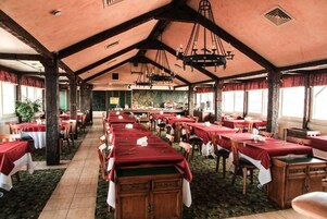 A large dining hall with long tables covered in red tablecloths, wooden chairs, and a high ceiling with exposed beams and chandeliers.