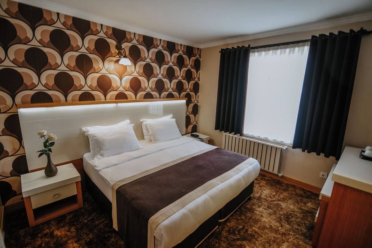 Una habitación de hotel con papel tapiz estampado, una cama con ropa de cama blanca y marrón, una mesita de noche con un jarrón de flores y una ventana con cortinas.